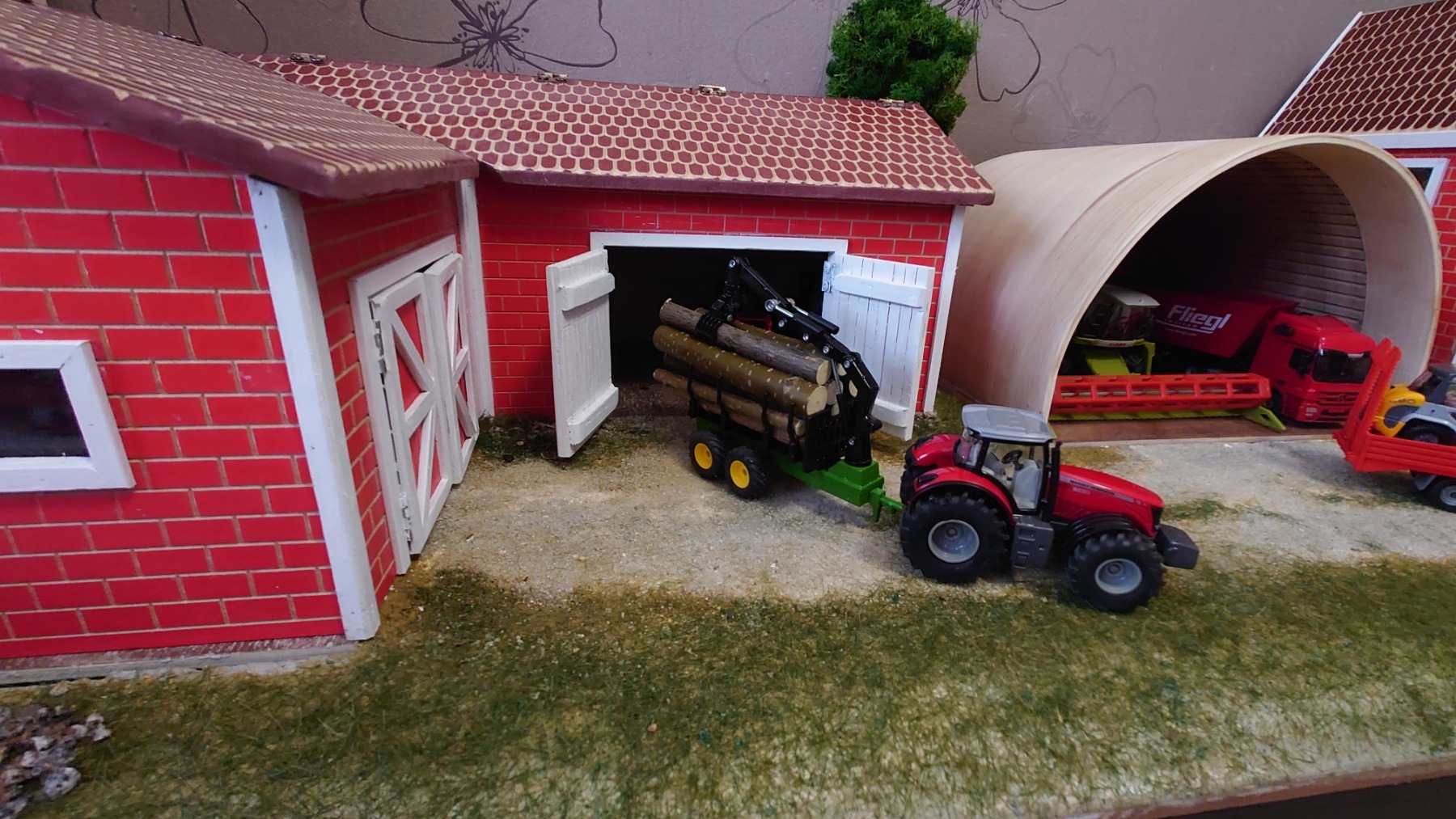Miniaturowa farma z czerwonymi budynkami, traktorem i przyczepą z drewnem, ukazująca scenę rolniczą w skali modelarskiej.