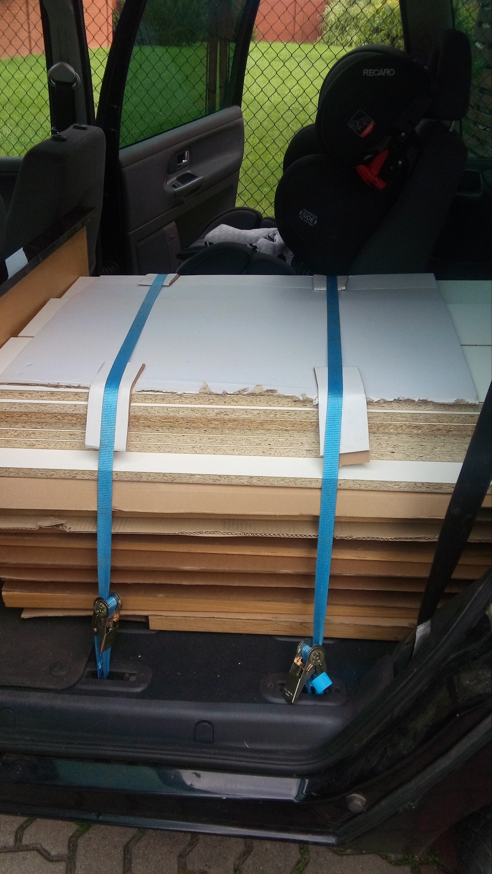 Paczka płyt meblowych zabezpieczona niebieskimi pasami transportowymi w bagażniku samochodu, widoczny fotelik dziecięcy Recaro.