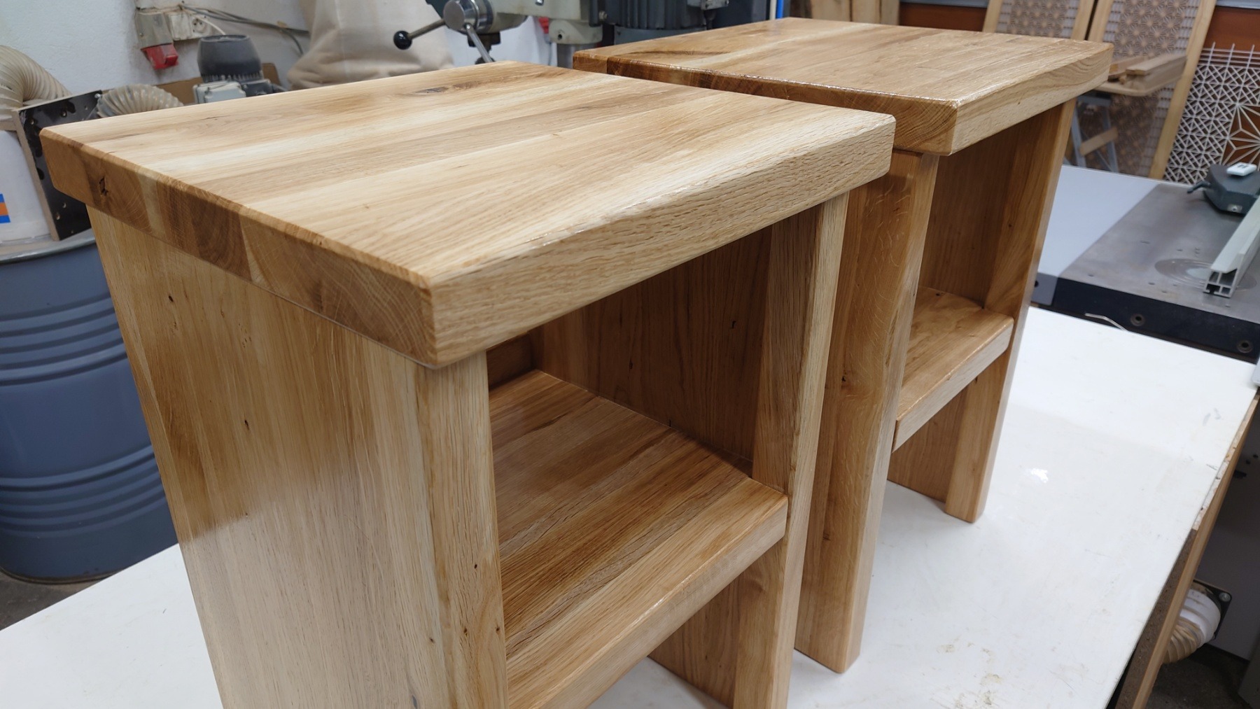 Dwa identyczne, jasne, dębowe stołki z półką, sfotografowane w warsztacie, z widocznymi śladami obróbki drewna i maszynami w tle.
