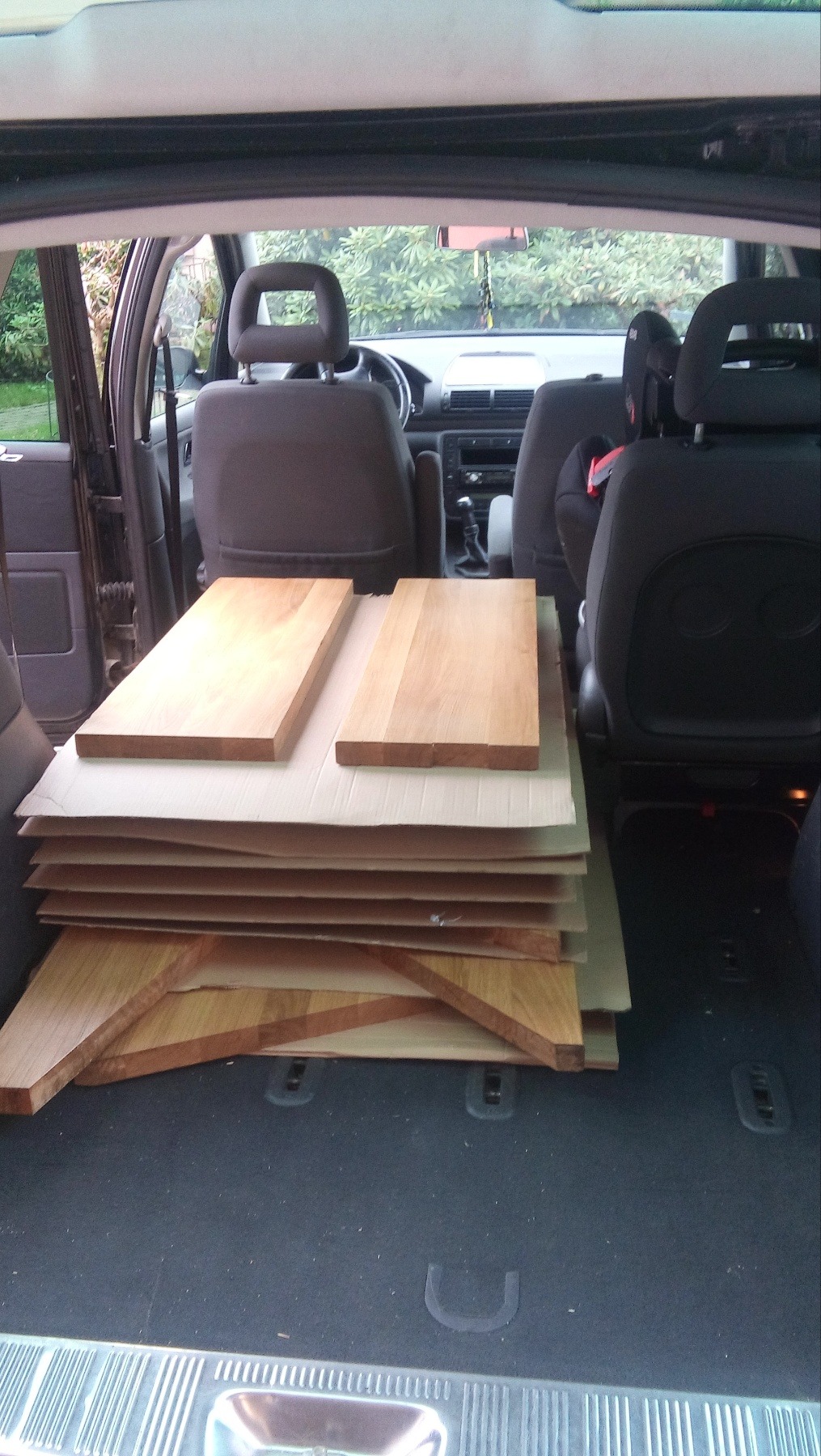 Deski drewniane ułożone na kartonie w bagażniku samochodu osobowego, widoczne fragmenty siedzisk i zagłówków foteli, tło z zielenią.