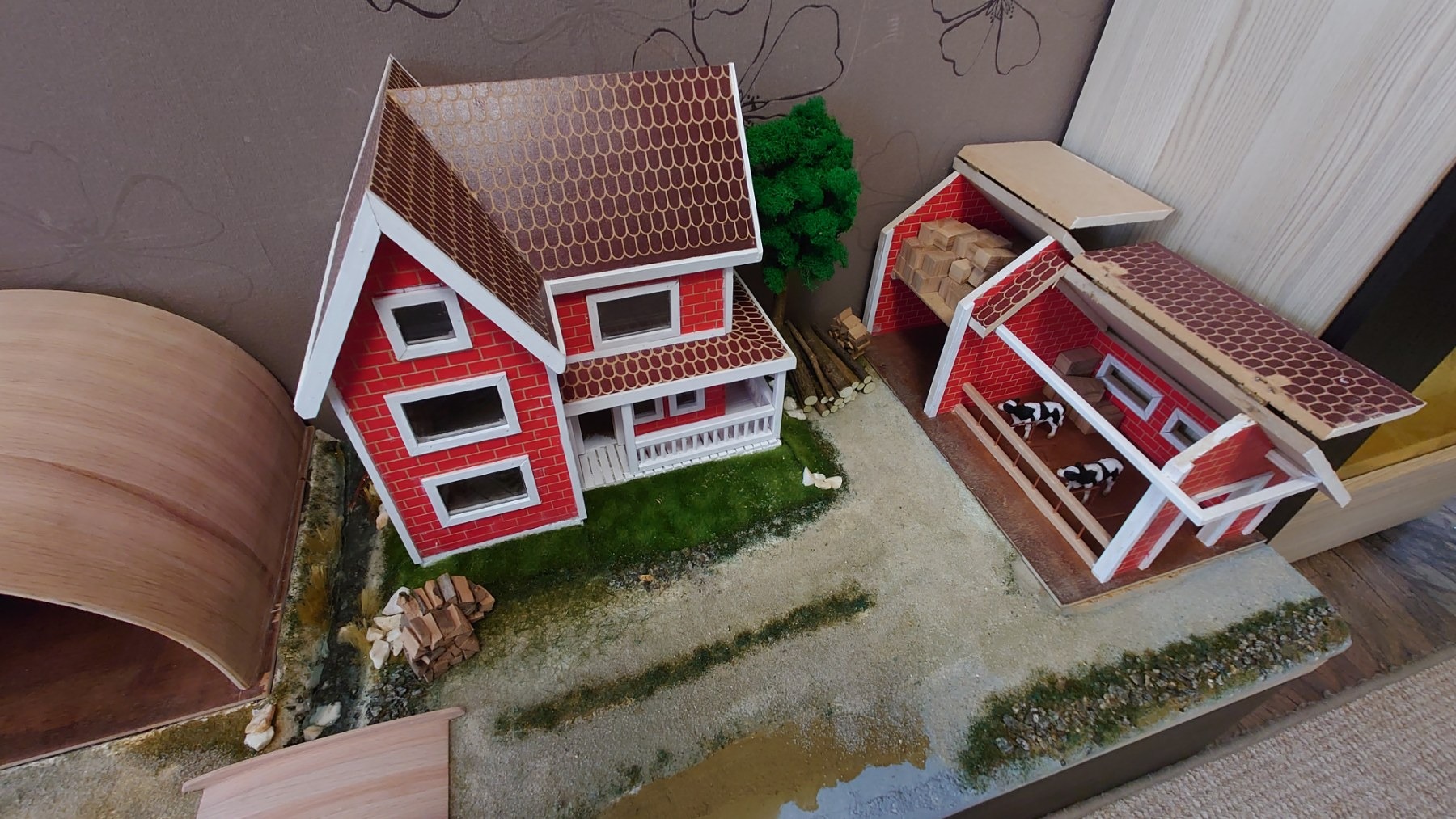 Makieta zagrody wiejskiej z czerwonym domem z białymi oknami, stodołą z krowami i stertą drewna, oraz elementami krajobrazu.