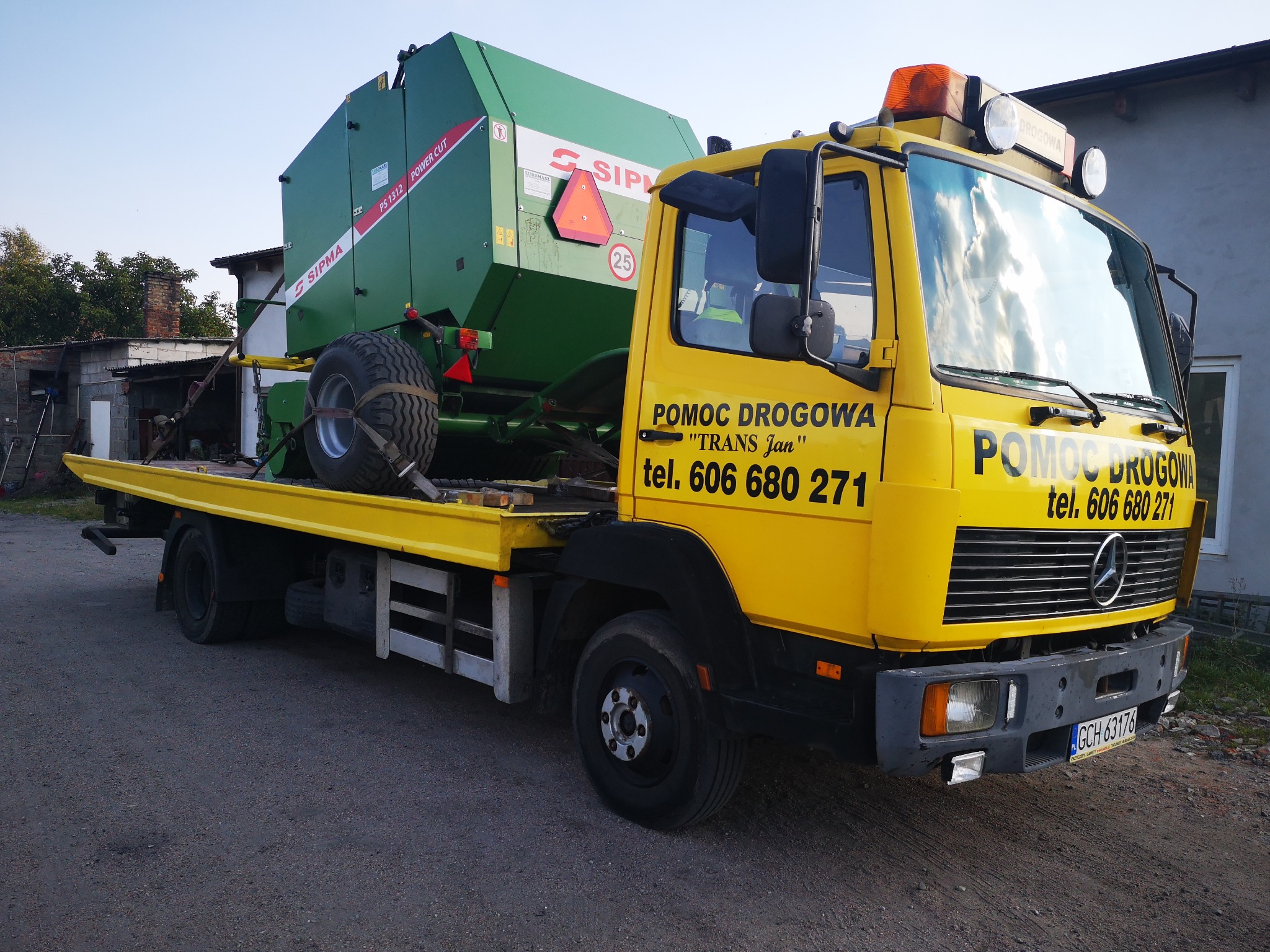 Żółta laweta pomocy drogowej 'TRANS Jan' przewozi zieloną prasę rolującą SIPMA PS 1312 Power Cut, widoczny numer telefonu kontaktowego na kabinie.