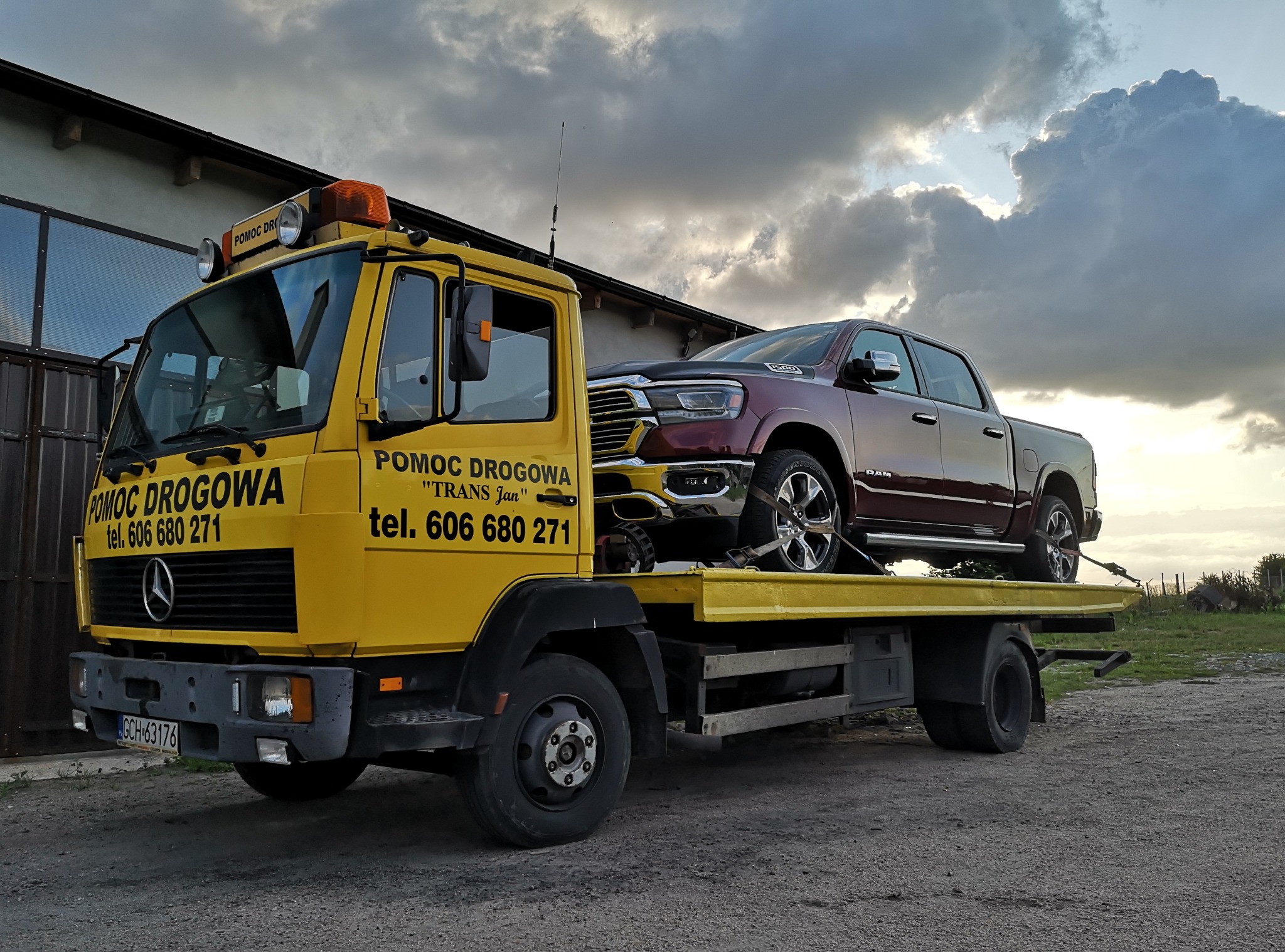 Żółta laweta z napisem 'Pomoc Drogowa' przewozi bordowego pickupa marki RAM, zabezpieczonego pasami transportowymi. W tle budynek i zachmurzone niebo.