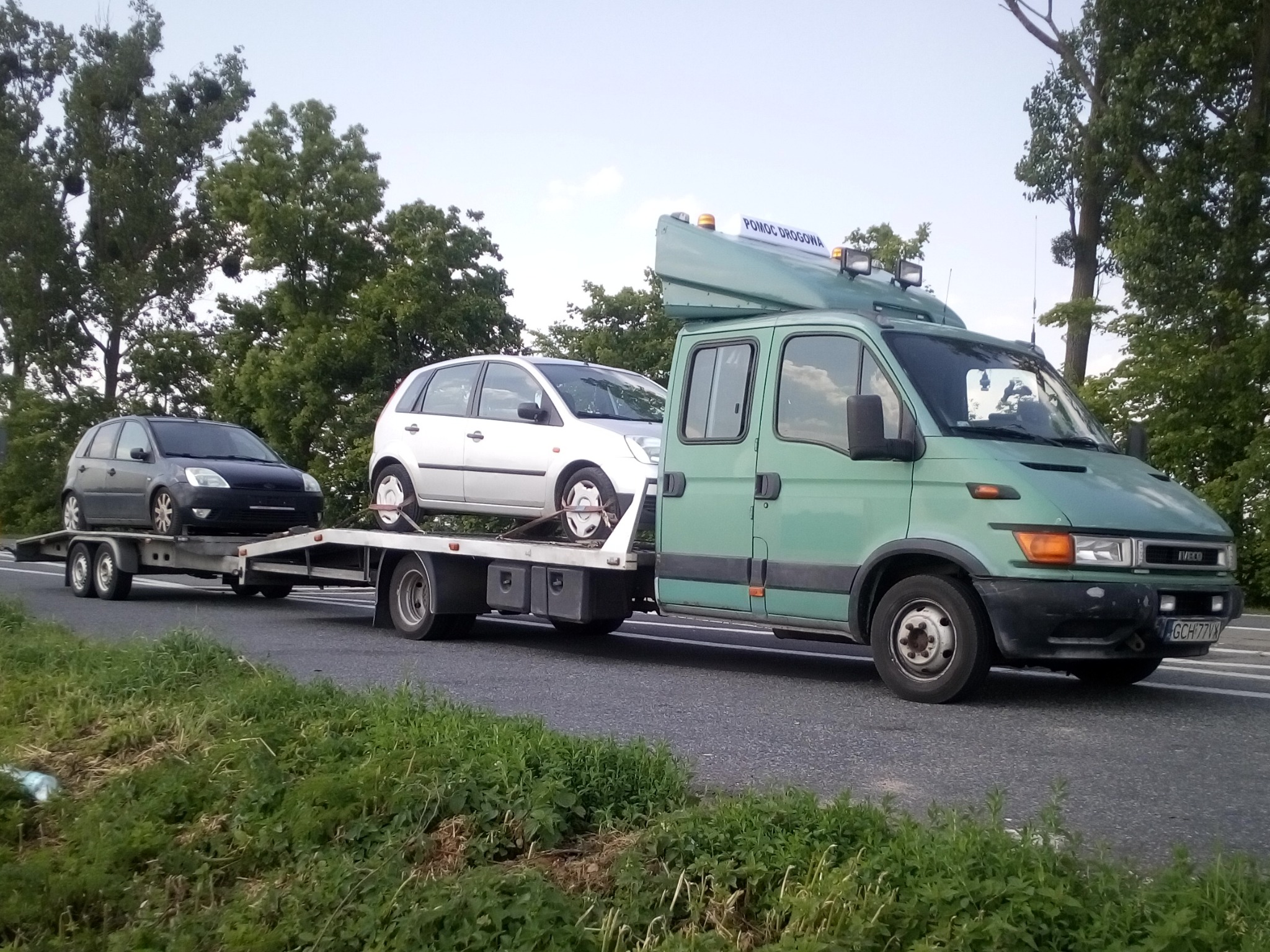 Zielona laweta z dwoma samochodami osobowymi na platformie, widok z boku na drodze, tablica 'Pomoc Drogowa' na dachu pojazdu.