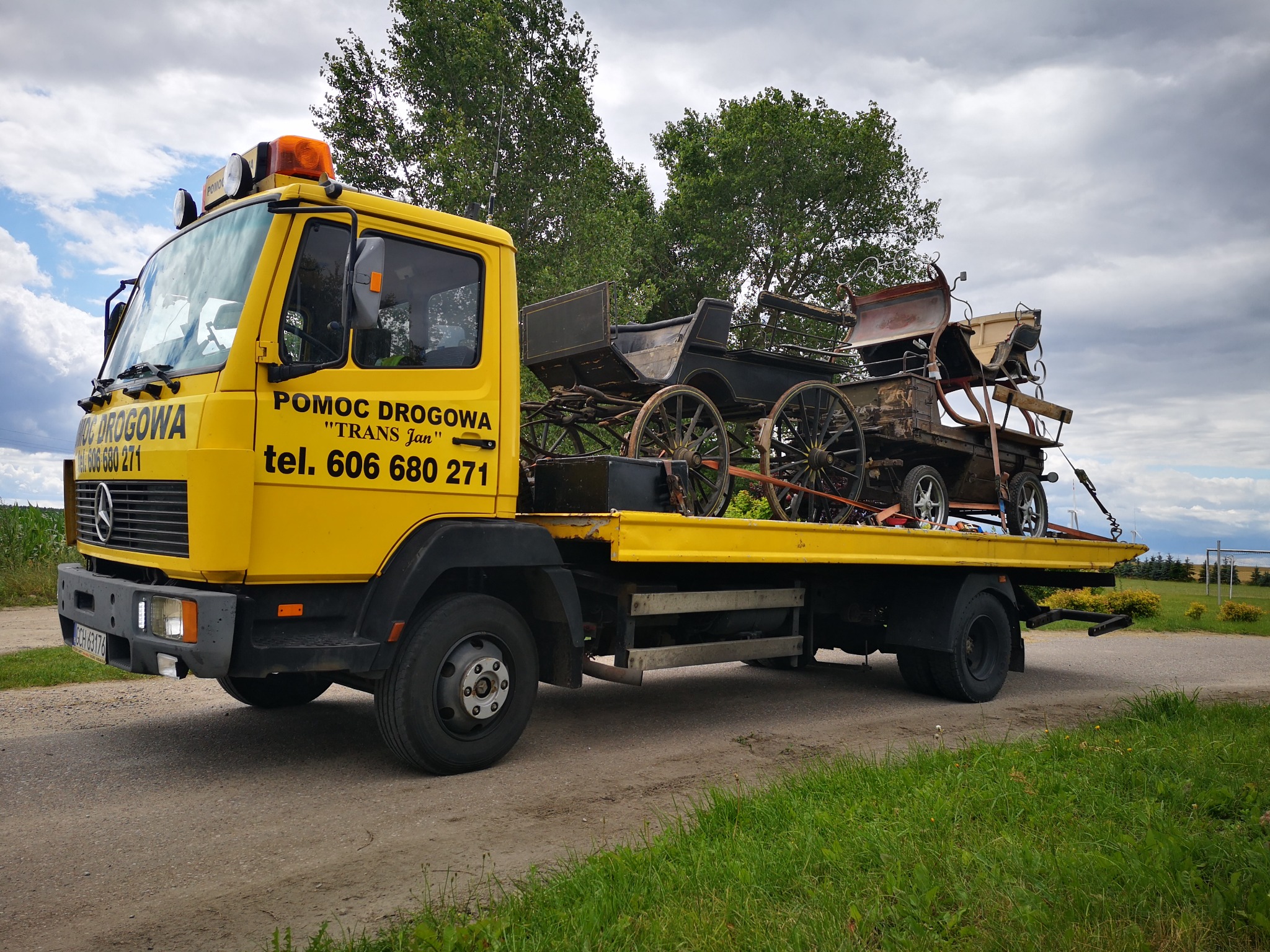 Żółta laweta z napisem 'POMOC DROGOWA TRANS Jan' przewozi dwie stare, drewniane bryczki na platformie, na tle pochmurnego nieba i zieleni