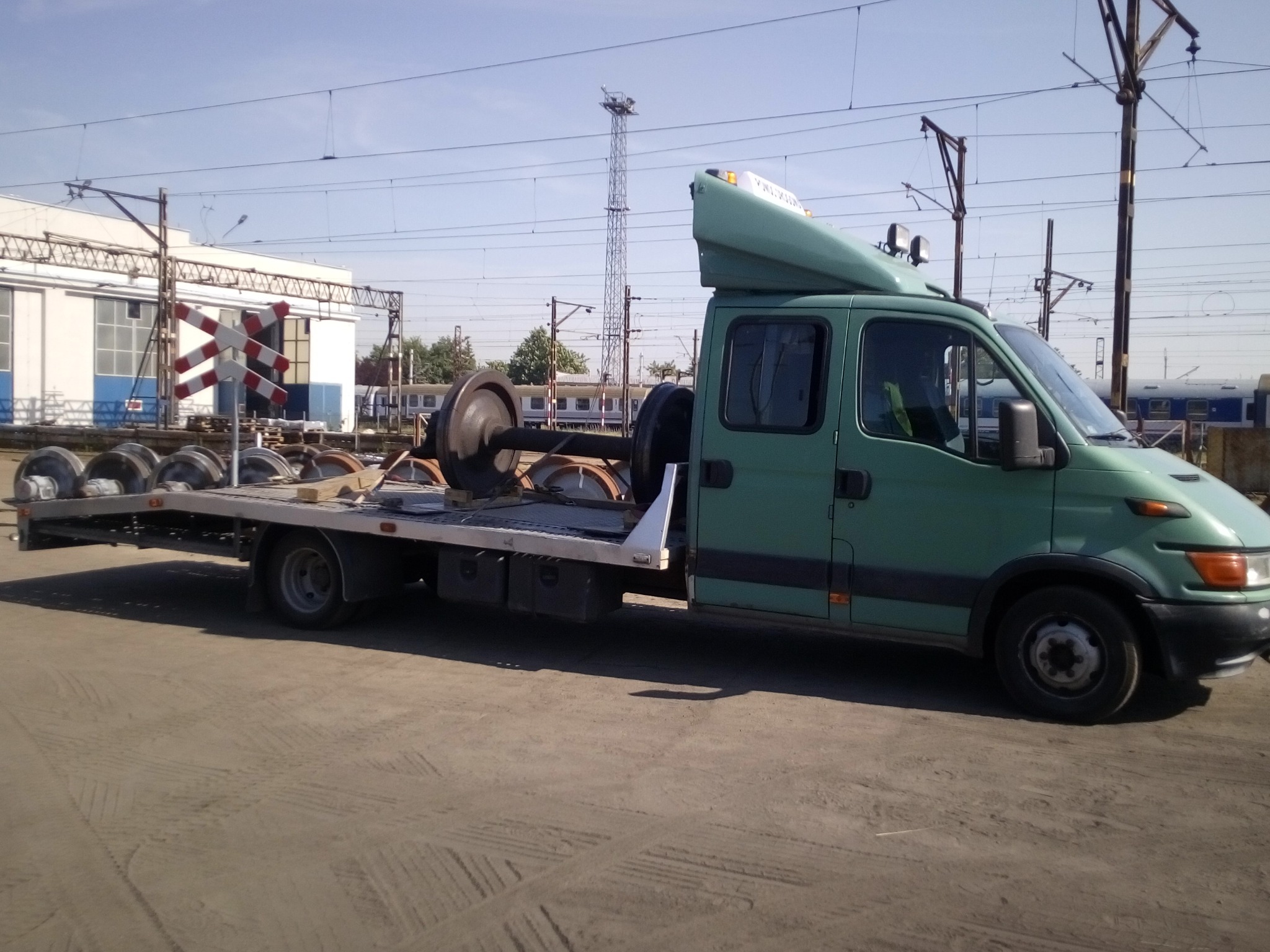 Zielona laweta z platformą transportową przewozi zestaw kół kolejowych na tle infrastruktury kolejowej i błękitnego nieba.