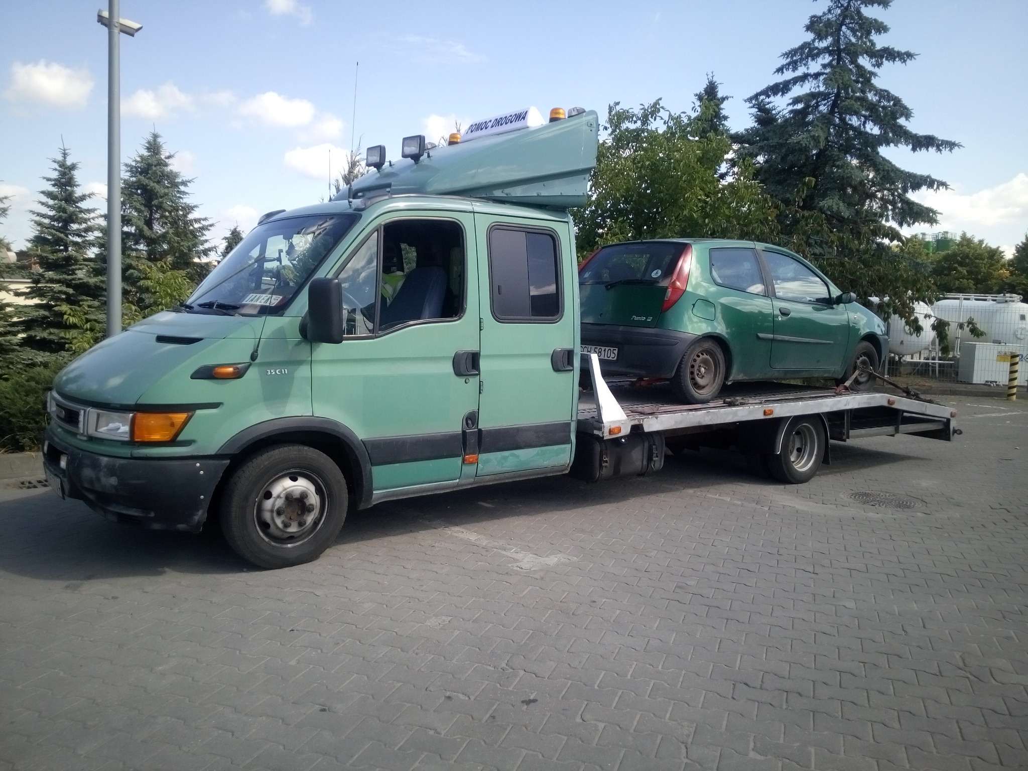 Laweta w Lichnowach z zielonym samochodem osobowym na platformie, zaparkowana na brukowanym placu w słoneczny dzień.