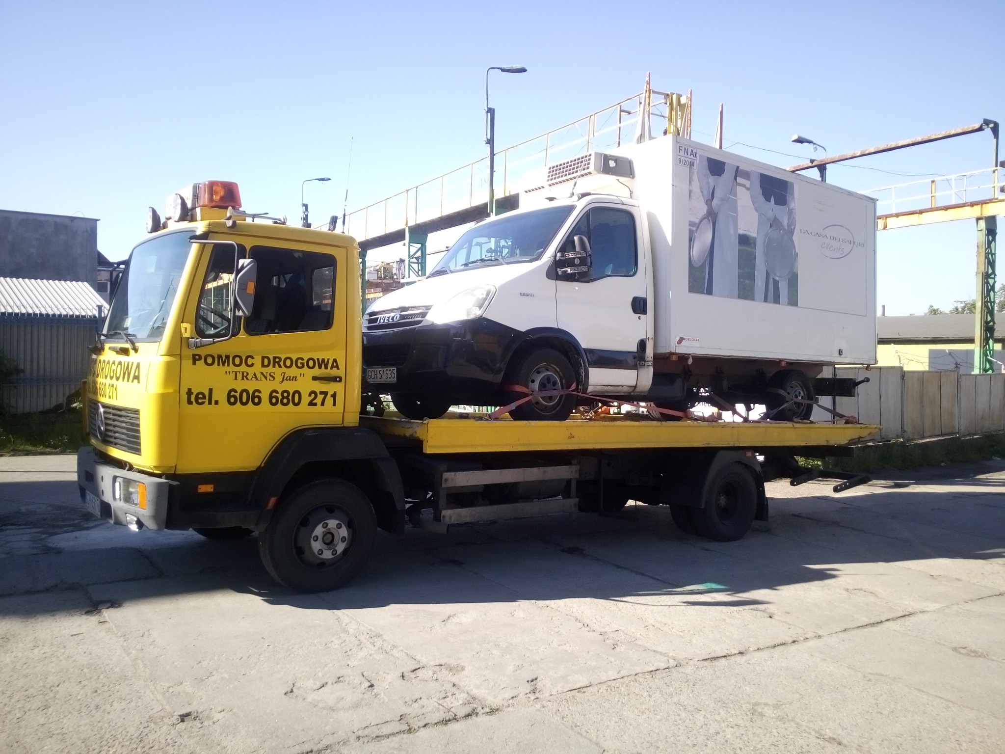 Żółta laweta z napisem 'POMOC DROGOWA TRANS Jan tel. 606 680 271' przewozi biały samochód dostawczy z reklamą cateringową na zabudowie.