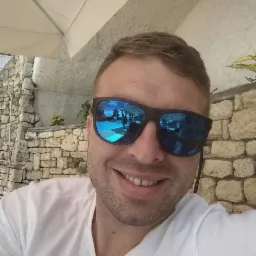 Uśmiechnięty mężczyzna w okularach przeciwsłonecznych z odbiciem basenu i kamiennej ściany w tle. Selfie w jasnym świetle.