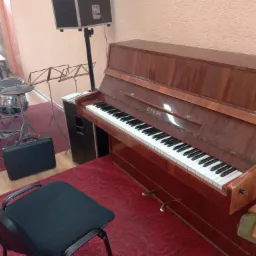 Brązowe pianino marki Etiuda w pokoju muzycznym z perkusją i czarnym krzesłem na czerwonym dywanie.