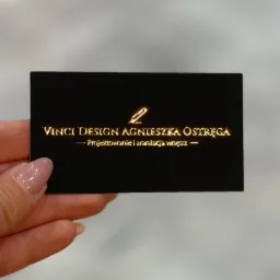 Czarna wizytówka z błyszczącym złotym napisem 'VINCI DESIGN AGNIESZKA OSTRĘGA - Projektowanie i aranżacja wnętrz' trzymana w dłoni z jasnoróżowym lakierem do paznokci.