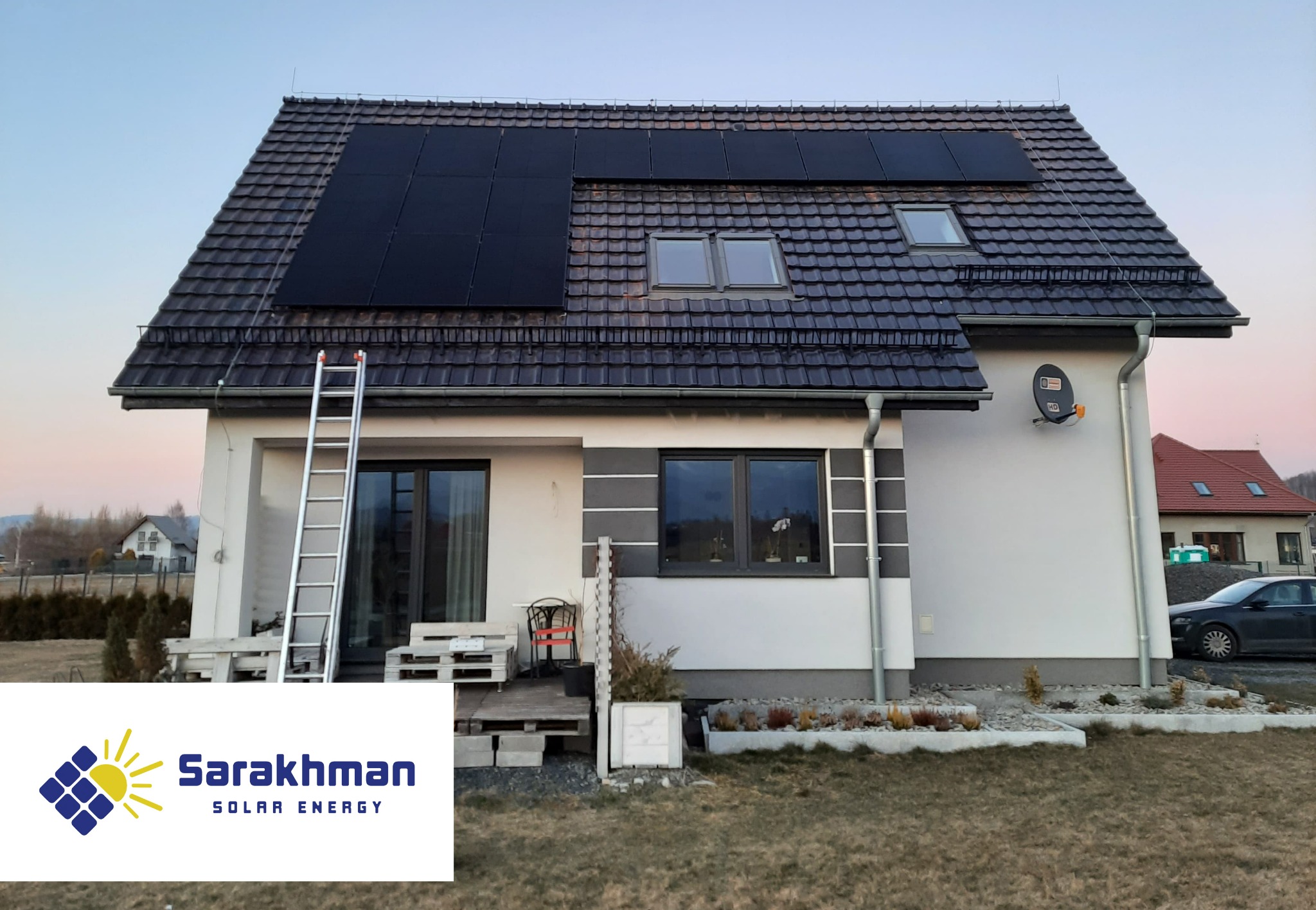 Dom jednorodzinny z panelami słonecznymi na dachu, drabina oparta o ścianę, widoczne logo firmy Sarakhman Solar Energy w dolnej części kadru.
