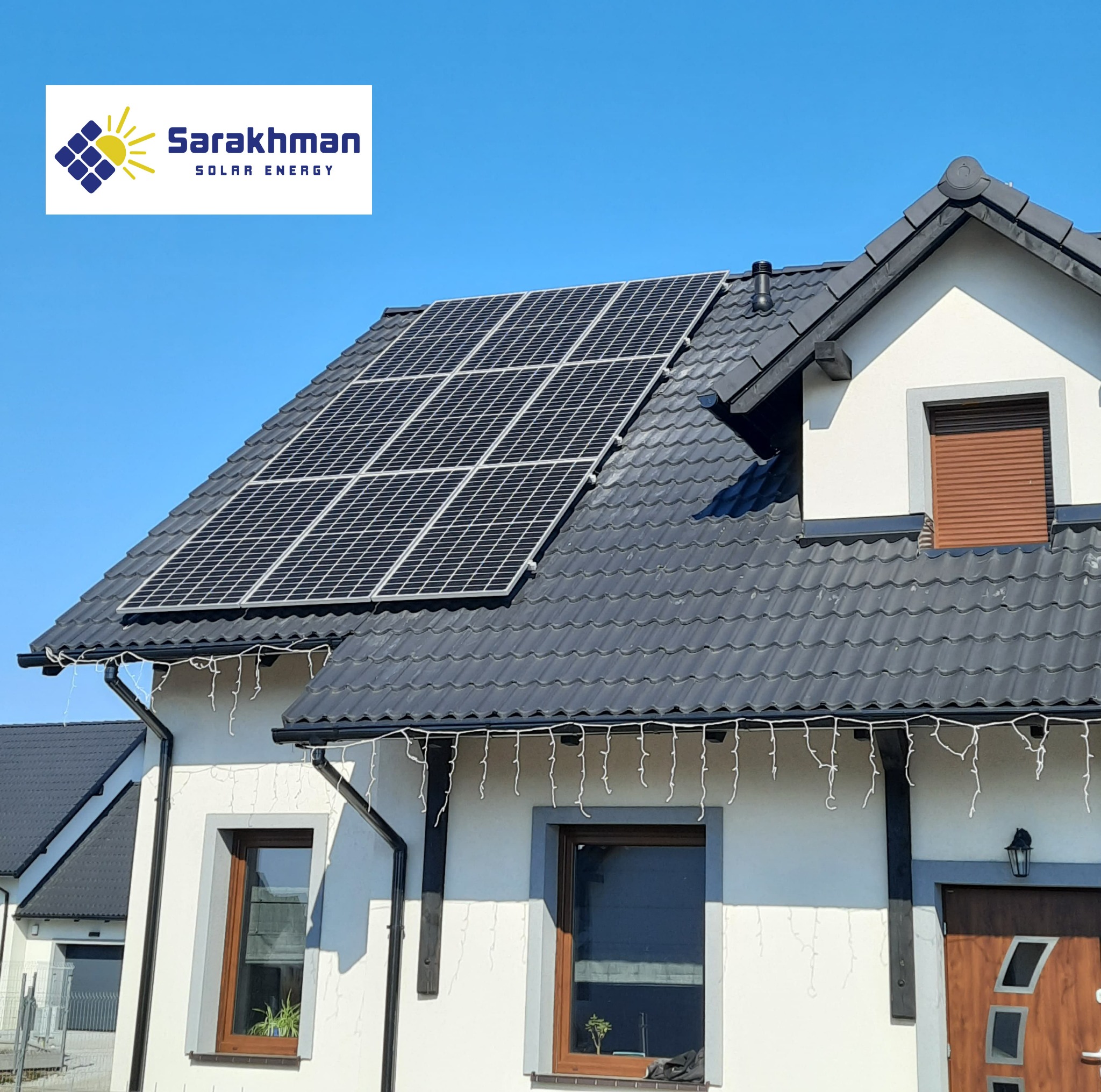 Panele fotowoltaiczne na dachu domu jednorodzinnego z ciemną dachówką, widoczne logo firmy instalującej panele Sarakhman Solar Energy, jasne niebo w tle.