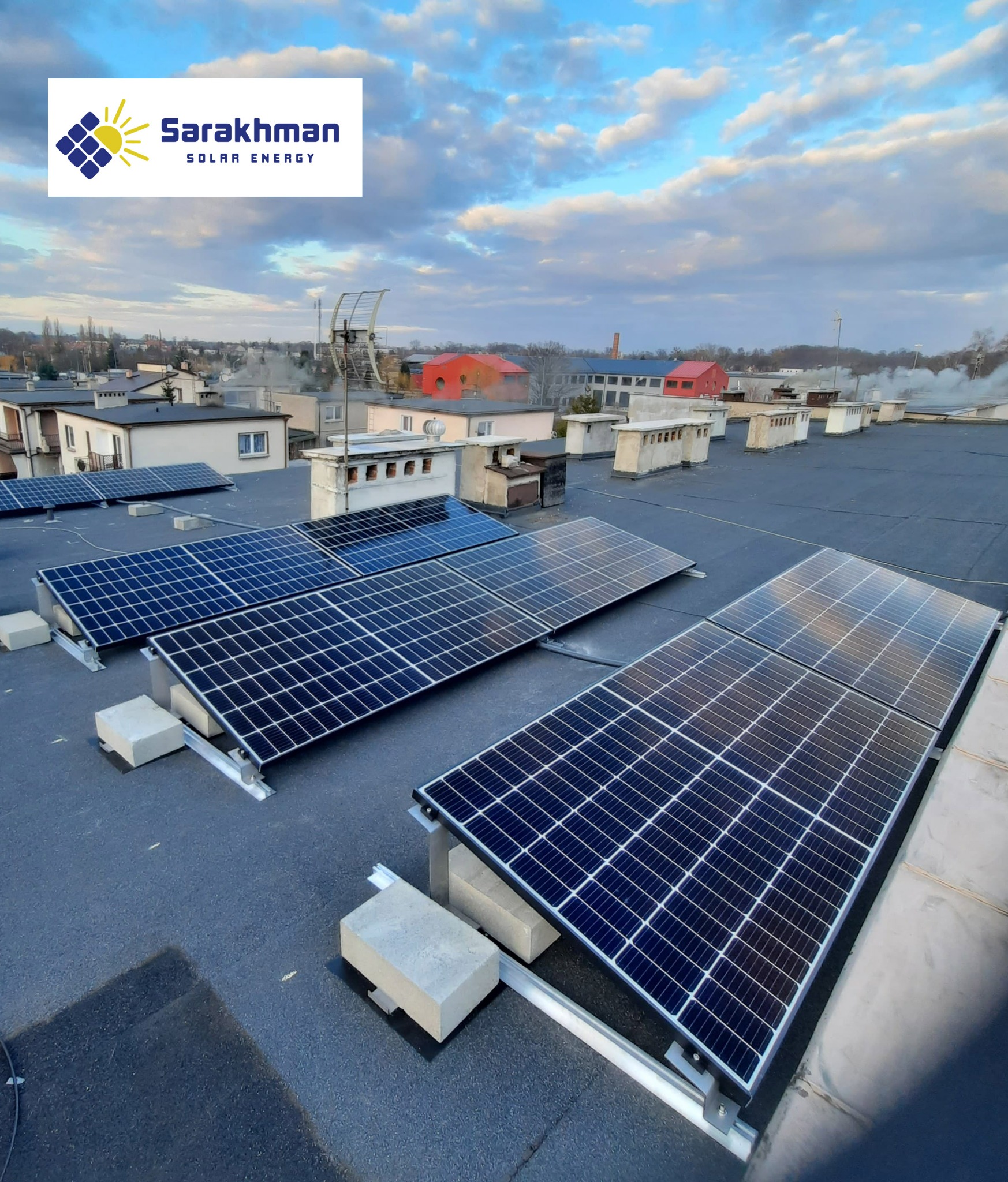 Panele fotowoltaiczne zamontowane na płaskim dachu budynku przemysłowego, widoczne betonowe bloczki stabilizujące konstrukcję, w tle zabudowa miejska i pochmurne niebo.