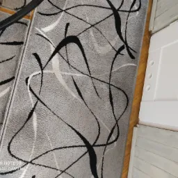 Szary dywan z abstrakcyjnym wzorem w trakcie czyszczenia odkurzaczem, widoczne stopy osoby obsługującej odkurzacz.