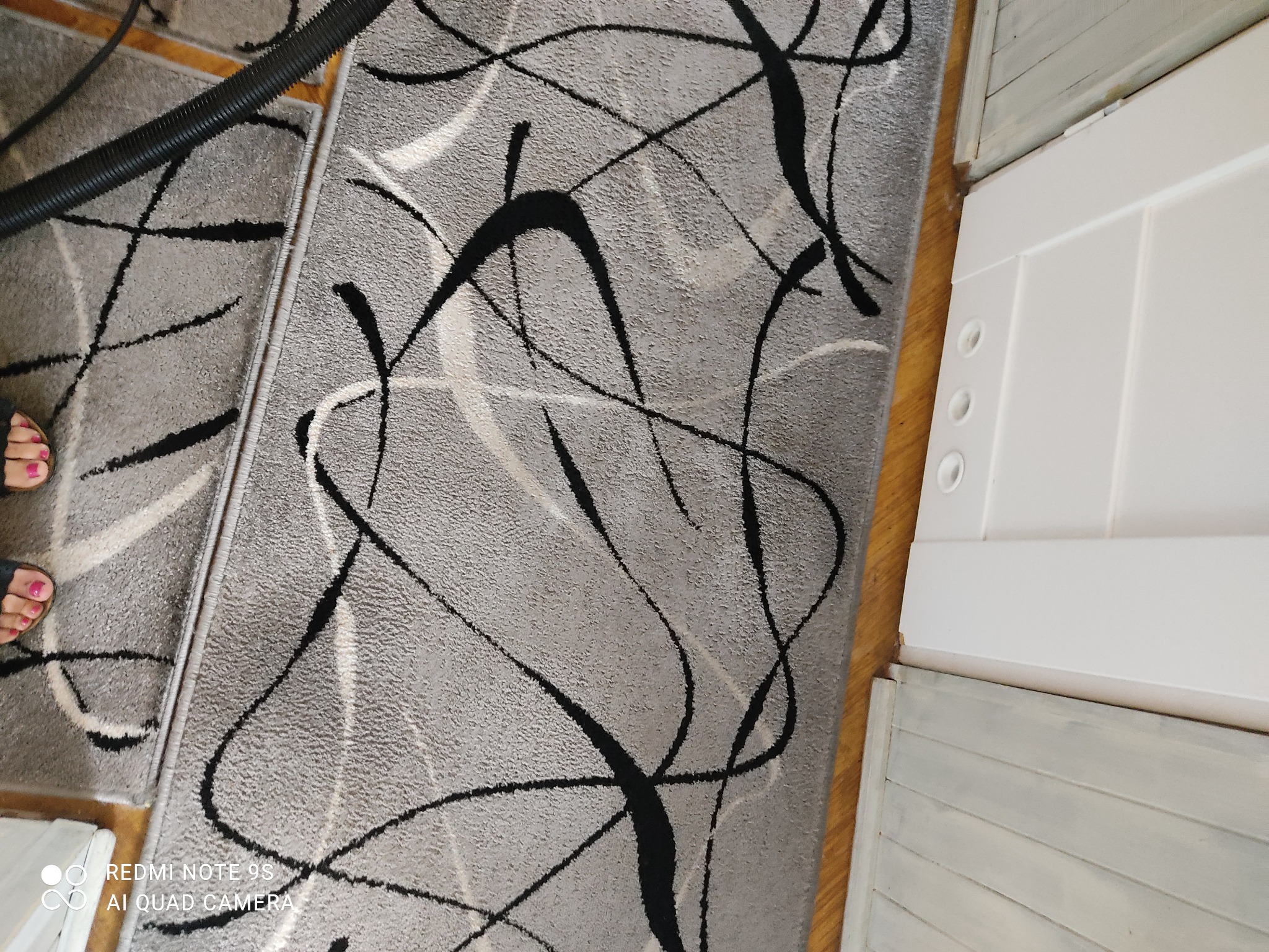 Szary dywan z abstrakcyjnym wzorem w trakcie czyszczenia odkurzaczem, widoczne stopy osoby obsługującej odkurzacz.