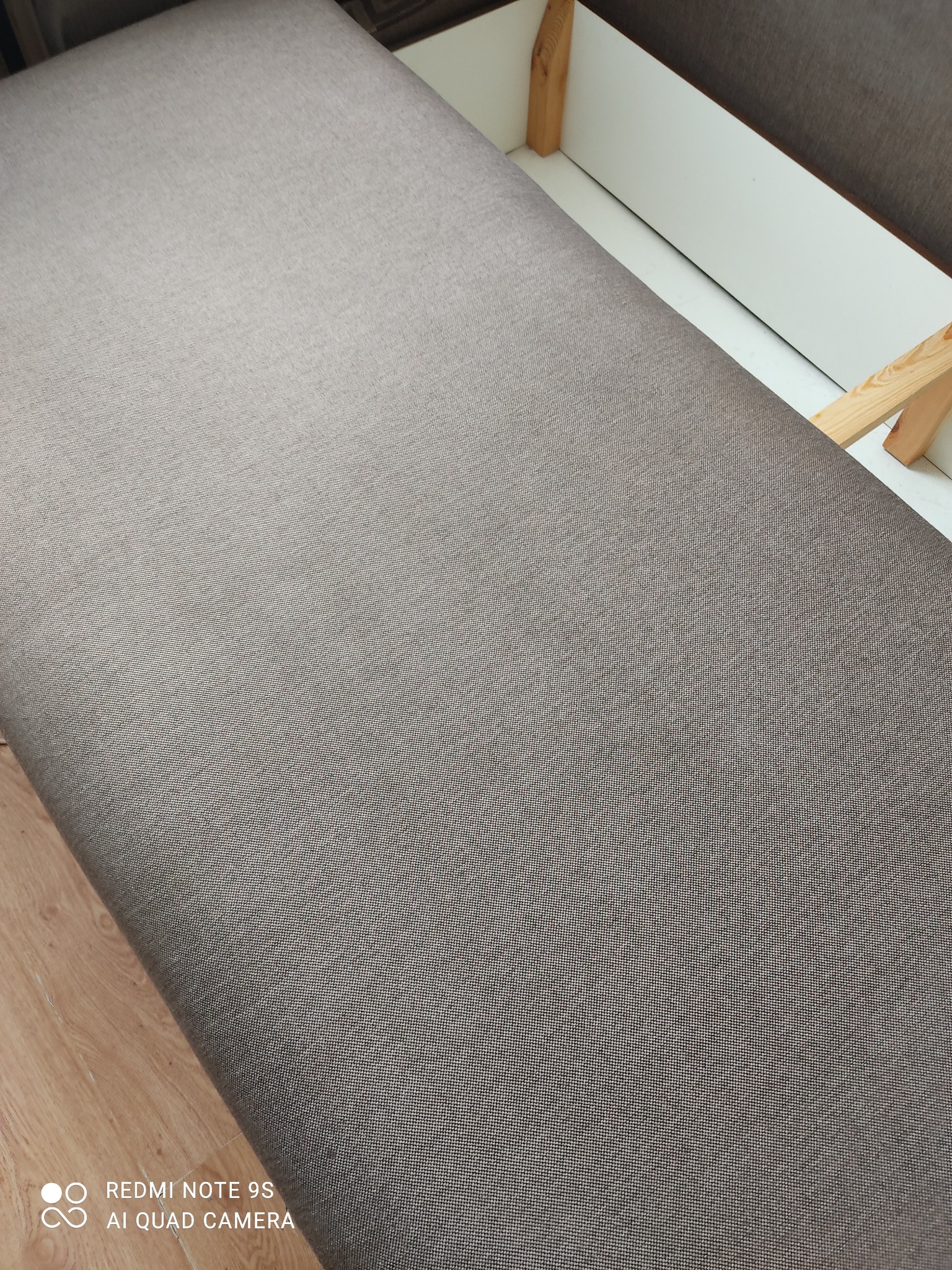 Szary materiał tapicerski na siedzisku mebla z widoczną drewnianą konstrukcją i białym wnętrzem skrzyni.