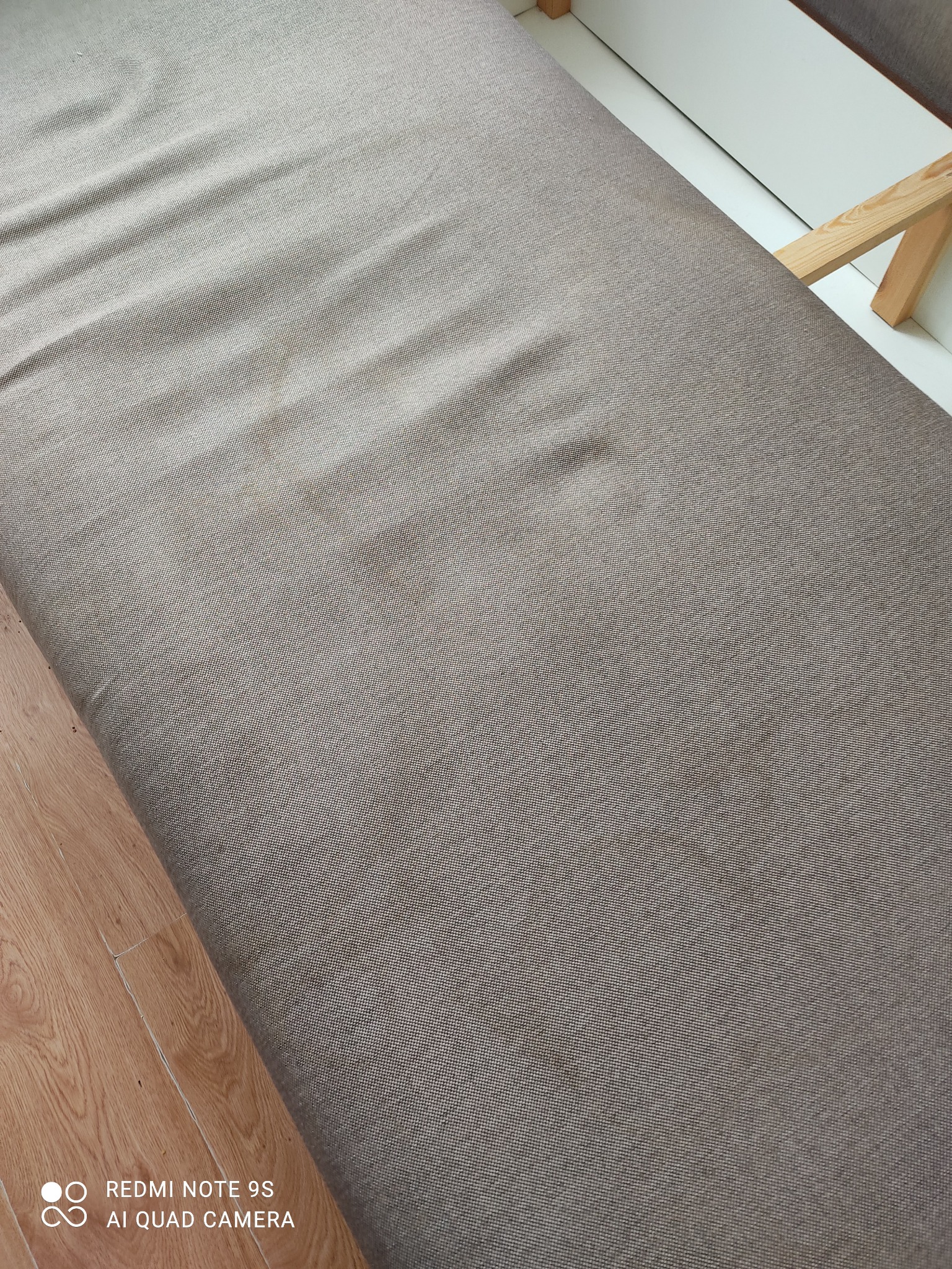 Szczegółowe ujęcie beżowej tkaniny obiciowej z widocznymi plamami na siedzisku kanapy lub tapczanu, z fragmentem drewnianej podłogi i białej ramy w tle.