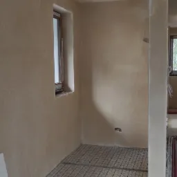 Wnętrze w trakcie prac wykończeniowych: świeżo położone tynki maszynowe na ścianach, widoczne okna z brązowymi ramami i instalacja ogrzewania podłogowego na podłodze. Część instalacji elektrycznej...