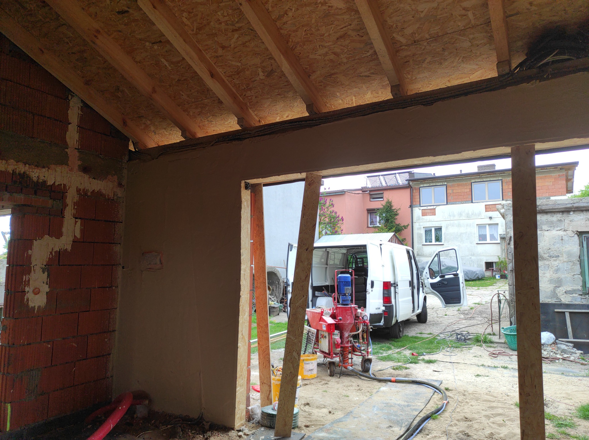 Wnętrze budynku w trakcie prac tynkarskich: ściany częściowo pokryte tynkiem, widoczna maszyna tynkarska na zewnątrz, biały bus z otwartymi drzwiami w tle.