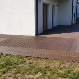 Nowoczesny taras z desek kompozytowych w kolorze brązowym, przylegający do białego budynku z oknami, otoczony trawnikiem.