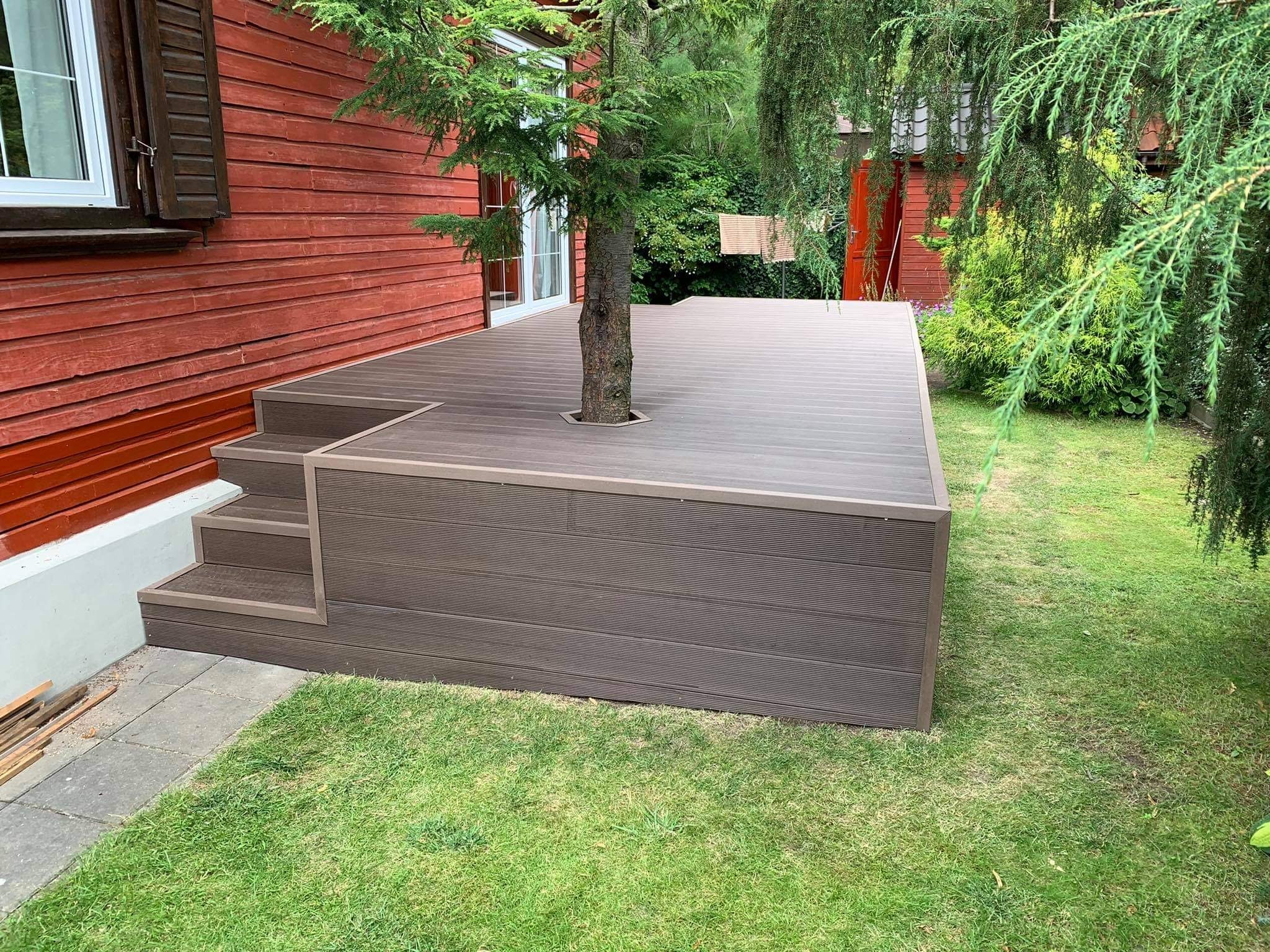 Nowoczesny, brązowy podest z desek kompozytowych otaczający rosnące drzewo, zintegrowany ze schodami przy czerwonym, drewnianym domu, otoczony zielonym trawnikiem.