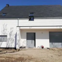 Nowoczesny, dwukondygnacyjny dom z płaskim dachem pokrytym szarą dachówką, białą elewacją, szarymi drzwiami garażowymi i wejściowymi oraz oknami z roletami, częściowo otoczony zielenią.