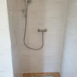 Kabina prysznicowa z chromowaną armaturą, wyłożona jasnymi płytkami imitującymi marmur, z podłogą z płytek drewnopodobnych i odpływem liniowym