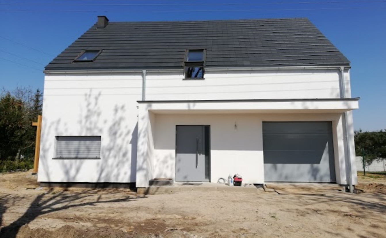 Nowoczesny, dwukondygnacyjny dom z płaskim dachem pokrytym szarą dachówką, białą elewacją, szarymi drzwiami garażowymi i wejściowymi oraz oknami z roletami, częściowo otoczony zielenią.