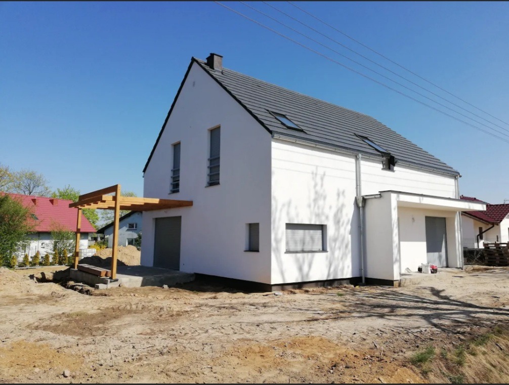 Nowoczesny dom jednorodzinny z szarą dachówką i jasną elewacją, widoczna drewniana konstrukcja zadaszenia tarasu w trakcie budowy, teren wokół domu w stanie surowym.