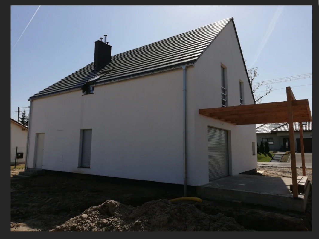 Nowoczesny dom jednorodzinny z białym tynkiem, grafitową dachówką i drewnianą pergolą nad wjazdem do garażu, widok z zewnątrz w słoneczny dzień.