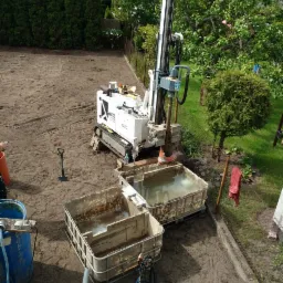 Mobilna wiertnica do studni głębinowych na działce, obok dwa zbiorniki z wodą, widoczne ślady wiercenia w ziemi.