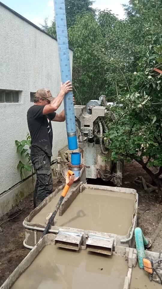 Pracownik łączy segmenty niebieskiego przewodu wiertniczego przy maszynie wiertniczej, w tle budynek i drzewa, widoczne zbiorniki z szlamem i pomarańczowa łopata.