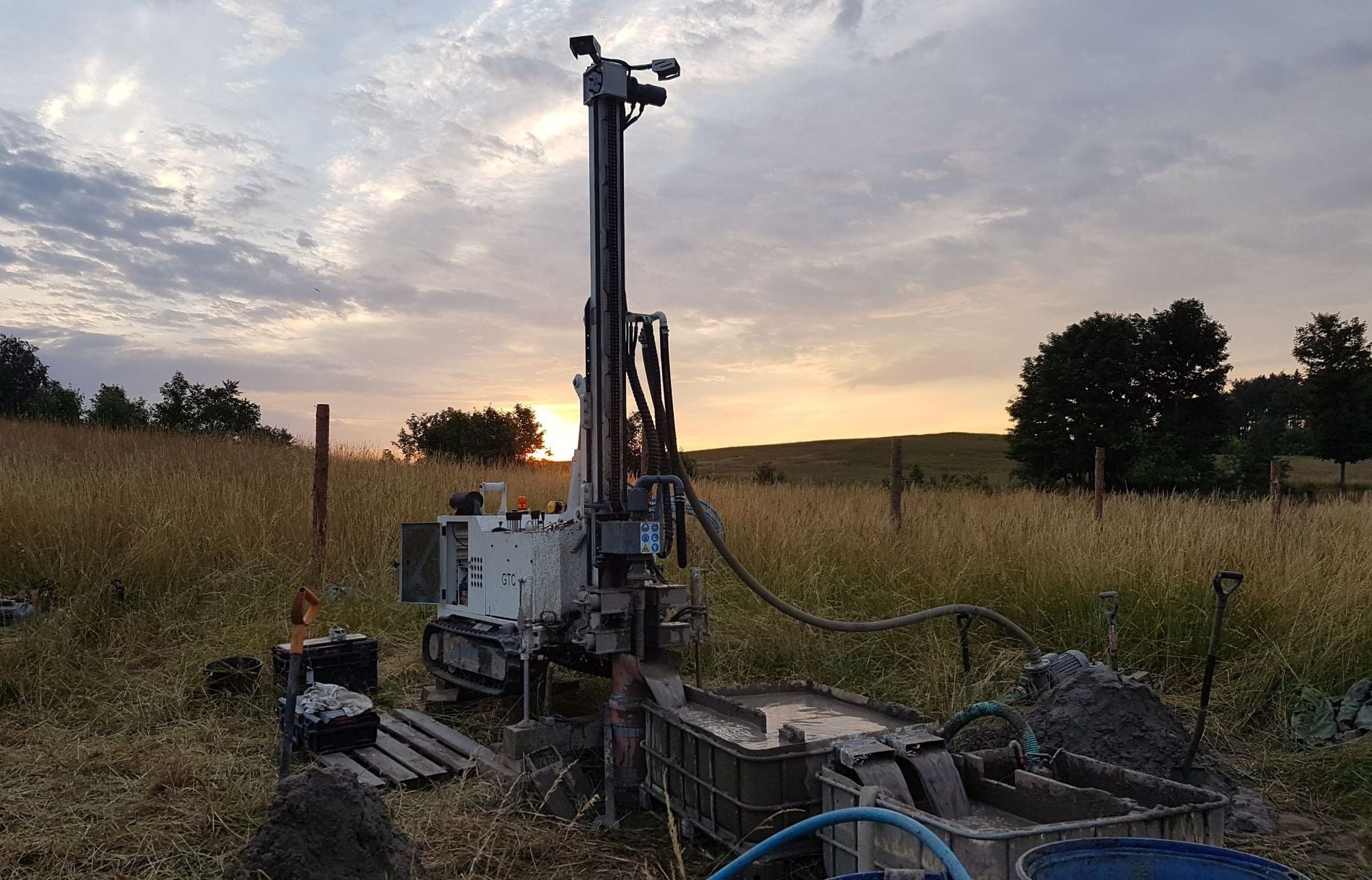 Wiertnica na gąsienicach podczas pracy przy zachodzącym słońcu, widoczne błoto i narzędzia, tło stanowią pola i drzewa.
