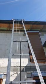 Długa, rozłożona aluminiowa drabina oparta o ścianę budynku z białych bloczków, prowadząca do dachu pod błękitnym niebem. Widoczne rolety zewnętrzne w kolorze brązowym.