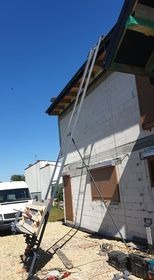 Długa aluminiowa drabina oparta o ścianę budynku w trakcie budowy dachu, na pierwszym planie stół roboczy z deskami, w tle biały bus i błękitne niebo.