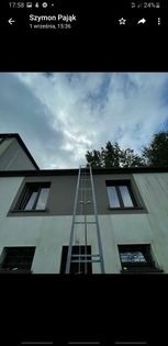 Długa, metalowa drabina przystawiona do dwupiętrowego budynku z szarą elewacją, widok z dołu na pochmurne niebo.