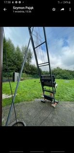 Czarna, metalowa konstrukcja przypominająca drabinę z podestem, ustawiona na zewnątrz, prawdopodobnie do prac na wysokościach, widoczna na tle trawnika i drzew.