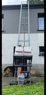 Wysoki, metalowy podnośnik budowlany ustawiony na trawniku przed budynkiem, widoczny pies rasy owczarek niemiecki w tle.