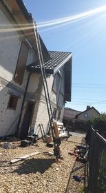 Podnośnik budowlany ustawiony przy elewacji domu w trakcie prac, widoczne materiały budowlane na ziemi, słoneczny dzień.