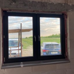 A2Group Sp. z.o.o - Czarne okno PCV z widokiem na podwórze z samochodem i furgonetką, zamontowane w surowym murze z betonu komórkowego, z widocznymi taśmami uszczelniającymi Soudal.