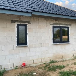 A2Group Sp. z.o.o - Surowy stan budynku z białego bloczka z zamontowanymi czarnymi oknami z białymi listwami, widoczne ślady po kleju montażowym, czerwone wiaderko przy ścianie, ziemia wokół budynku.