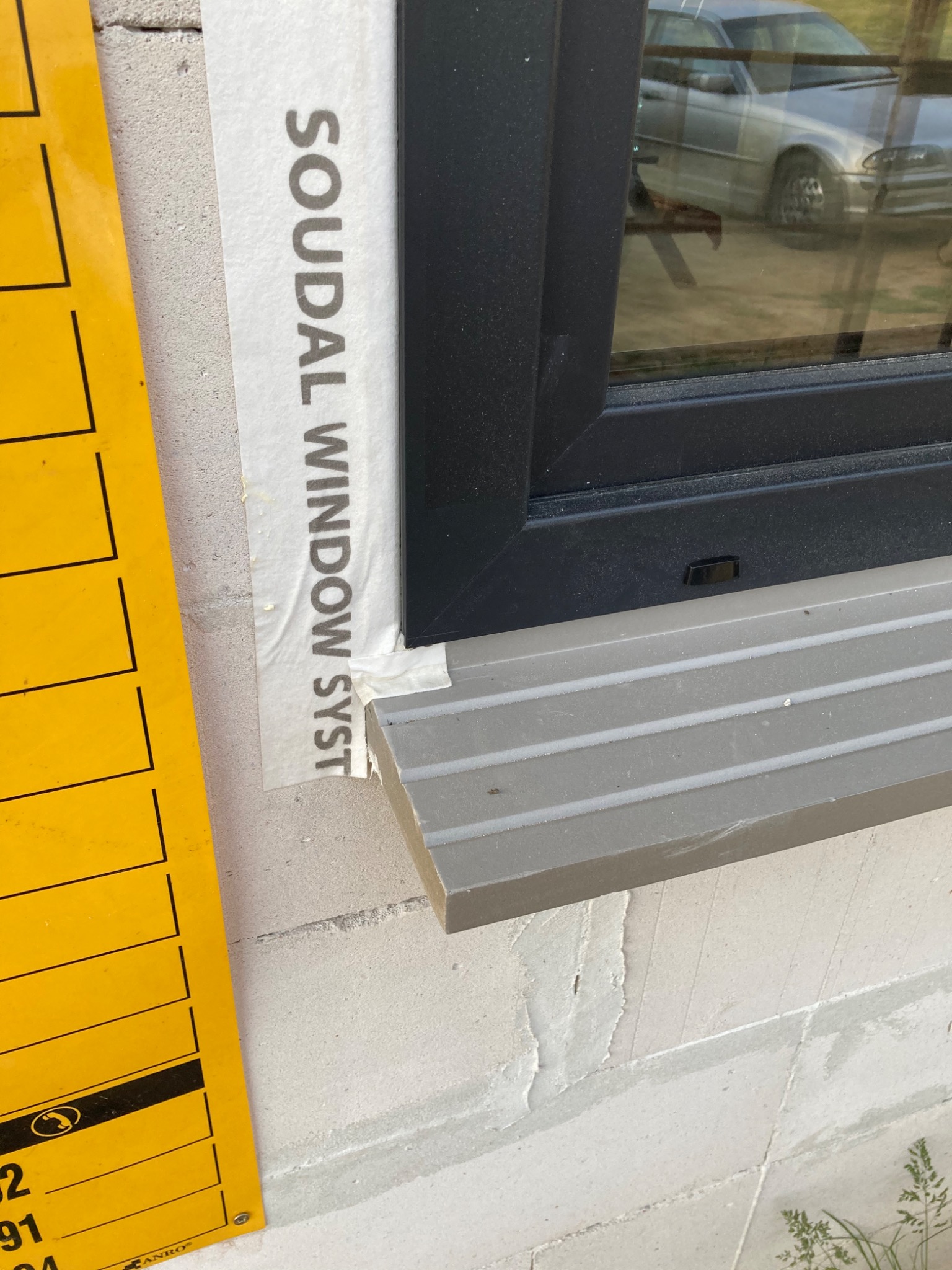 Fragment zamontowanego okna z czarną ramą i szarym parapetem zewnętrznym, zabezpieczone taśmą z napisem SOUDAL WINDOW SYSTEM, widoczny żółty element montażowy i odbicie samochodu w szybie.