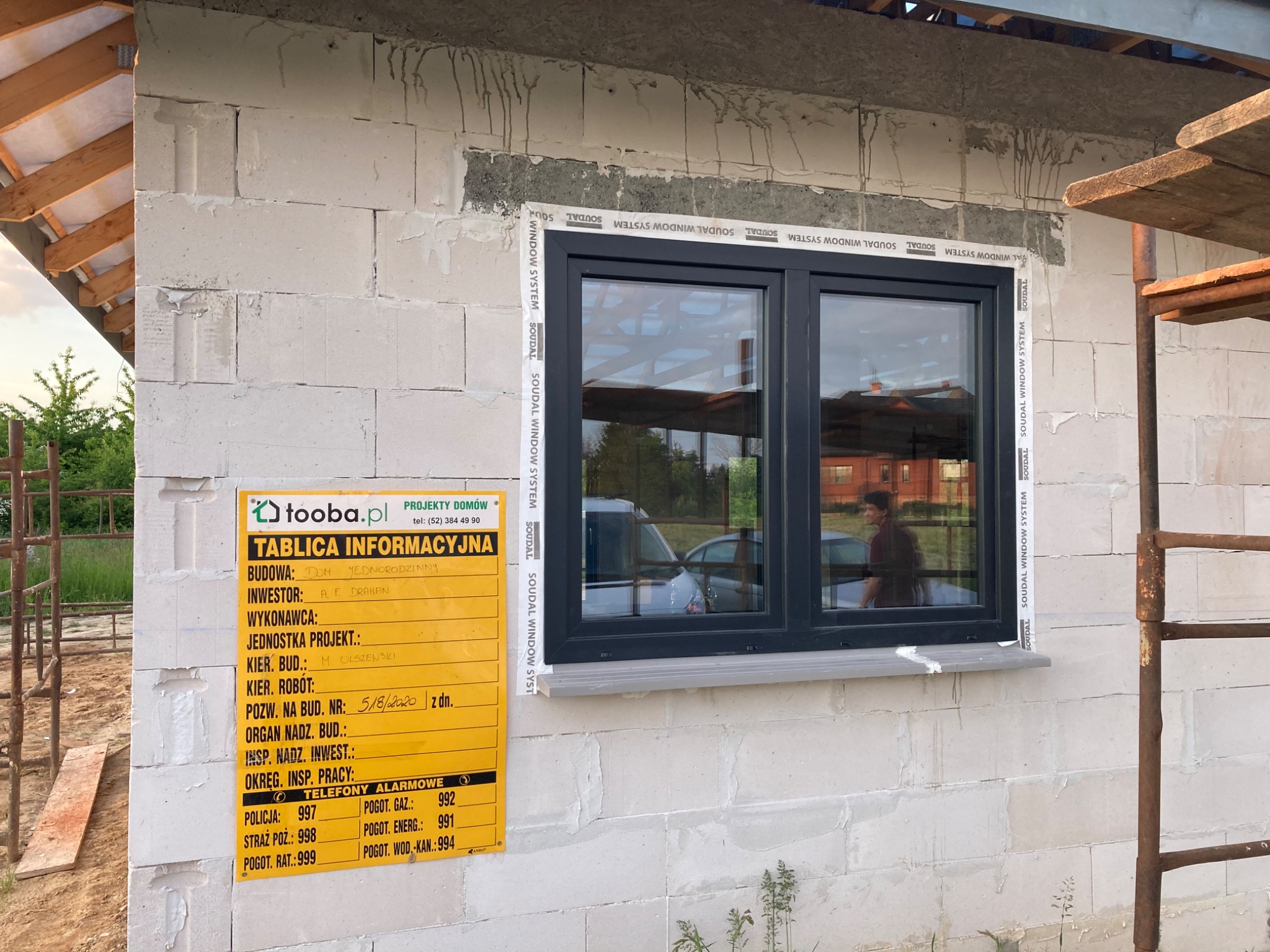 Nowe, czarne okno zamontowane w surowej ścianie z betonu komórkowego, zabezpieczone taśmą Soudal, z widoczną tablicą informacyjną budowy i fragmentem rusztowania.