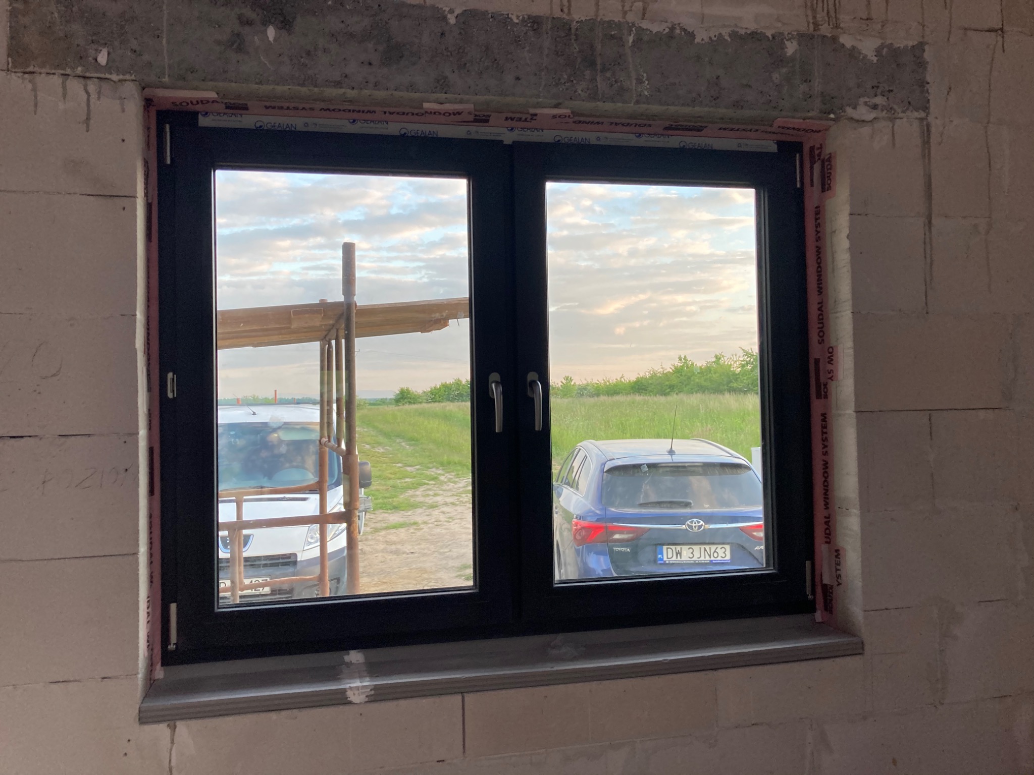 Czarne okno PCV z widokiem na podwórze z samochodem i furgonetką, zamontowane w surowym murze z betonu komórkowego, z widocznymi taśmami uszczelniającymi Soudal.