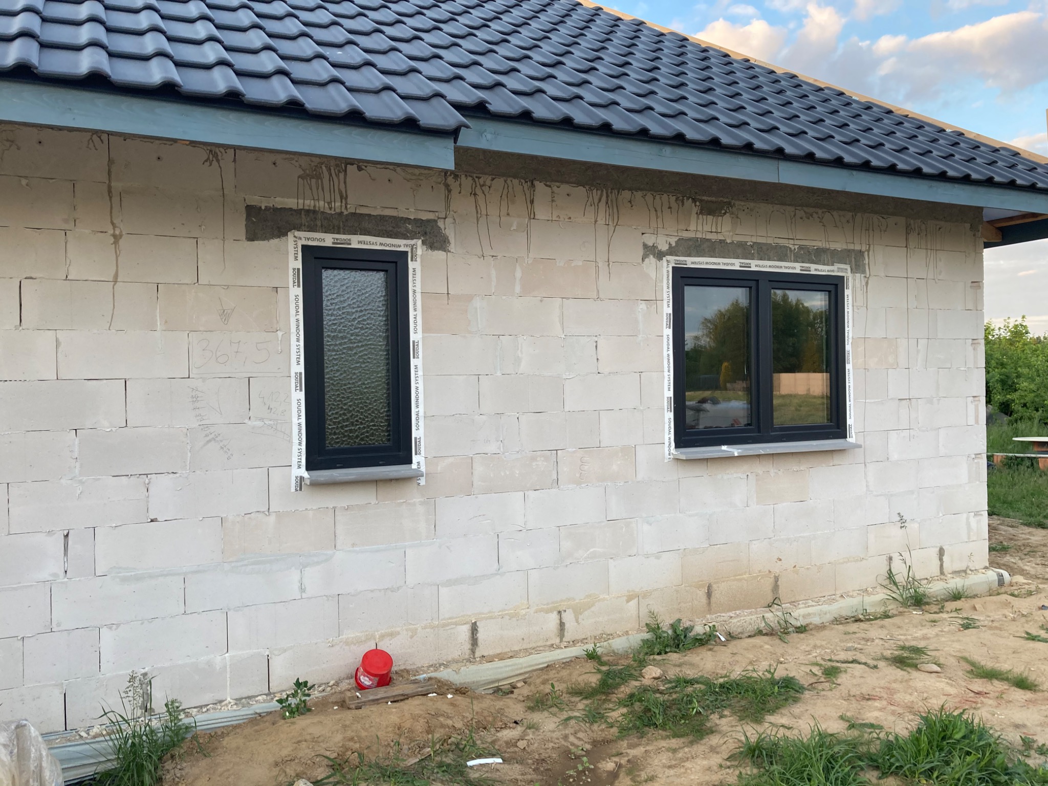 Surowy stan budynku z białego bloczka z zamontowanymi czarnymi oknami z białymi listwami, widoczne ślady po kleju montażowym, czerwone wiaderko przy ścianie, ziemia wokół budynku.