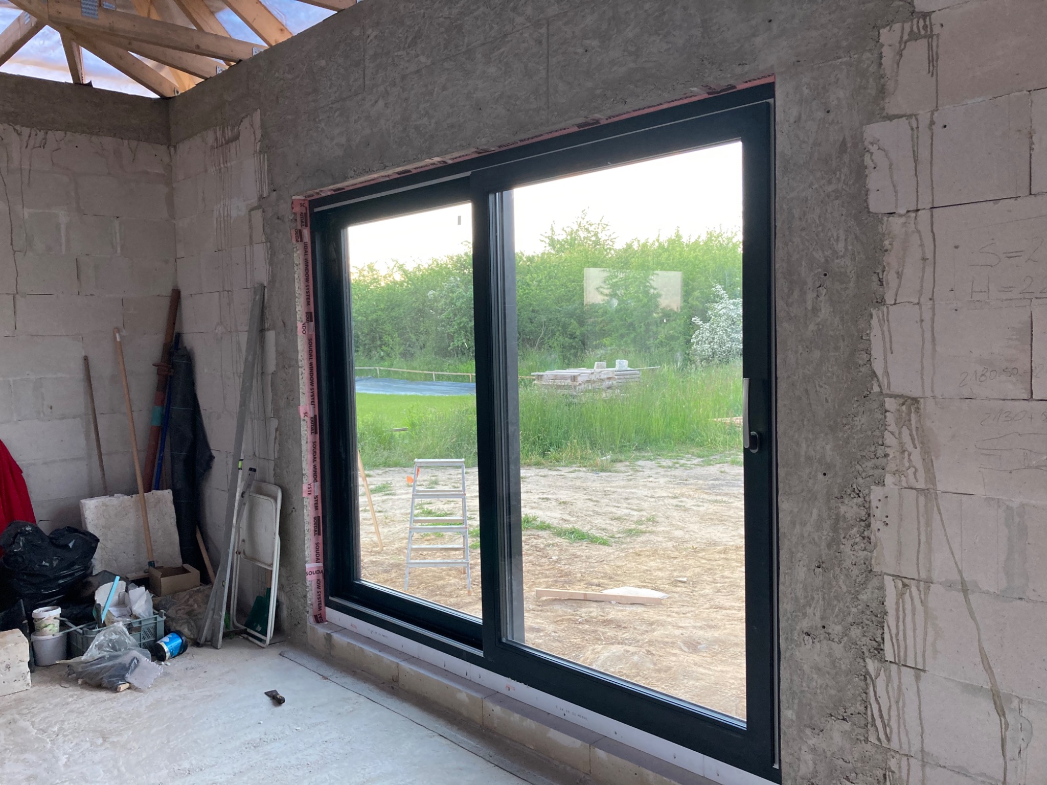 Duże przesuwne okno z czarną ramą, widok na zewnątrz na zieloną trawę i drzewa, ściany w surowym stanie, w tle drabina aluminiowa.