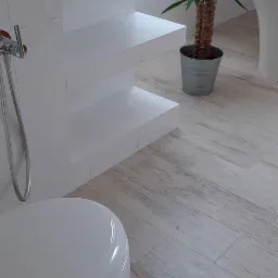 Nowoczesna łazienka z białymi płytkami imitującymi tapetę, drewnopodobną podłogą, wiszącą toaletą, półkami z płytek i rośliną w metalowej doniczce.