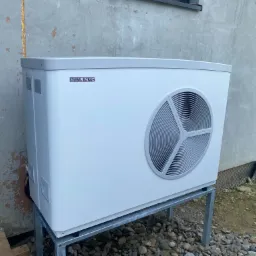 Biała pompa ciepła Stiebel Eltron zamontowana na metalowej konstrukcji przed budynkiem z szarą elewacją, widoczne okno w tle.