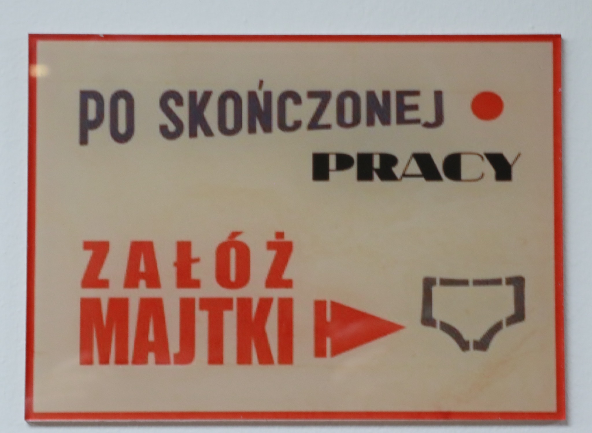 Tabliczka z humorystycznym napisem 'Po skończonej pracy załóż majtki' z symbolem majtek, umieszczona na ścianie.